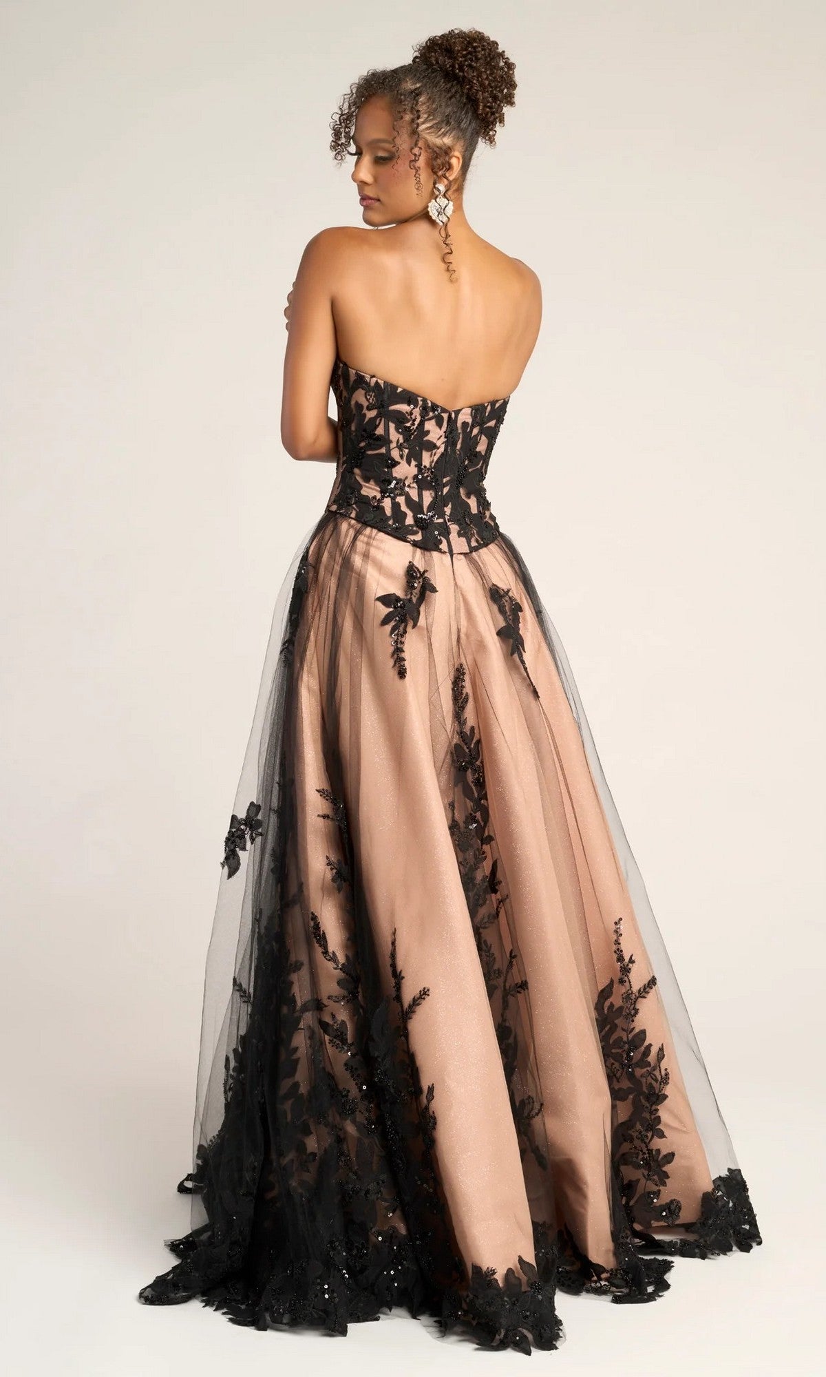 A woman stands with her back to the camera in the Portia and Scarlett PS26061 long formal dress, showcasing its strapless corset bodice, nude underlayer, and black lace overlay against a plain background.